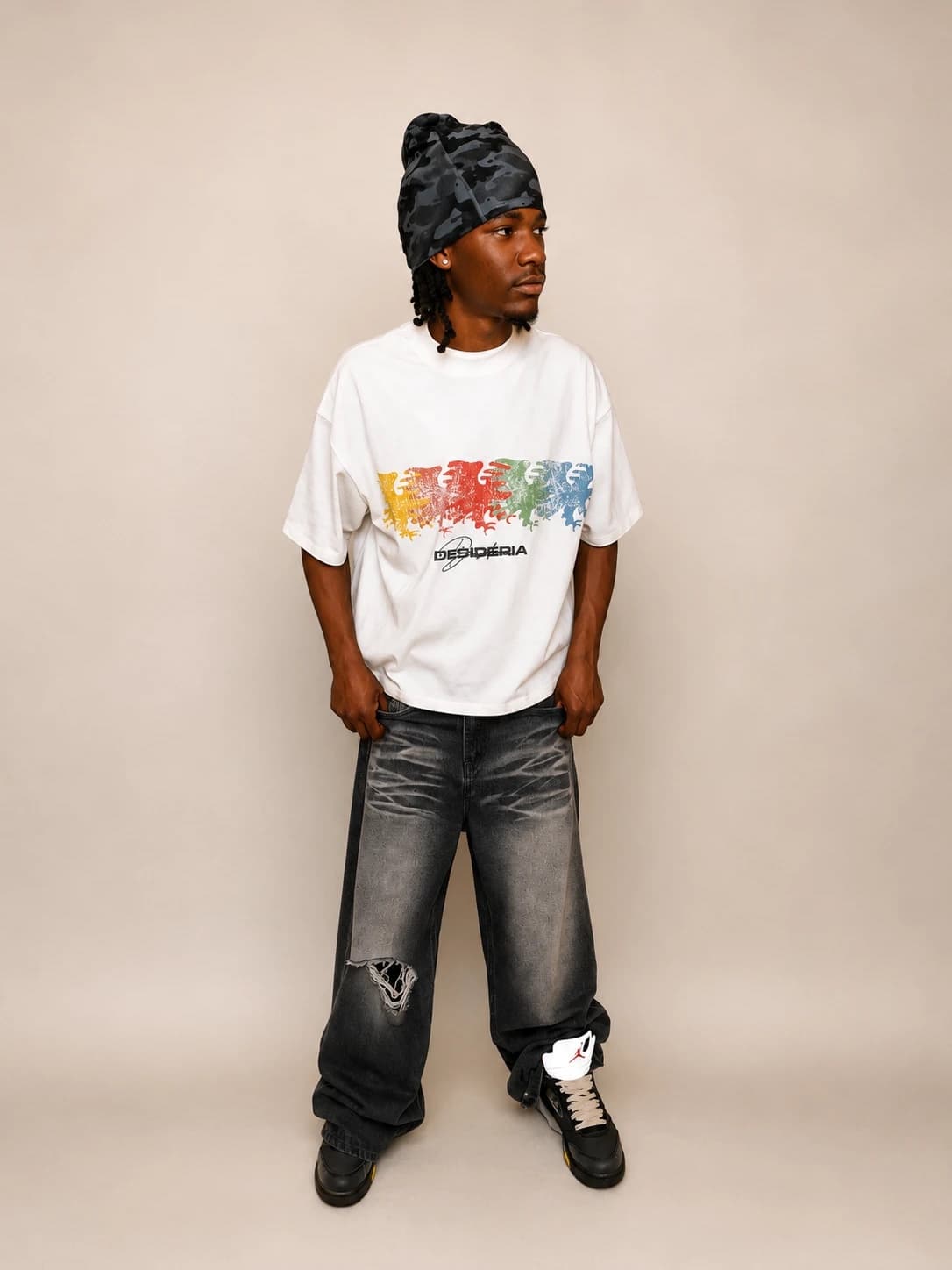 Model in the Dispersion tee and a camo beanie, shot in studio against a bone backdrop